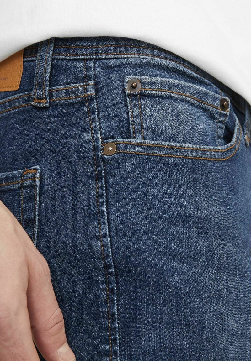 Jean en denim bleu foncé, avec un design standard à cinq poches, des rivets en cuivre et des coutures visibles le long des seams.