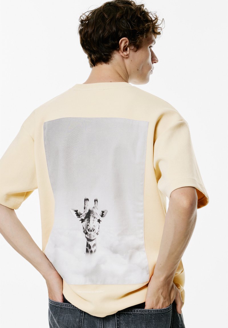 Gul bomulds t-shirt med korte ærmer, der har et stort gråt og sort grafisk motiv af et giraffehoved på ryggen, med subtil tekst nedenfor.