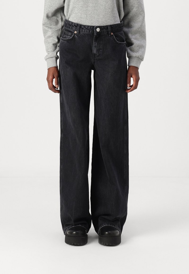 Personne portant un jean en denim noir ample, des chaussures noires et un haut gris à manches longues, debout devant un fond blanc uni.