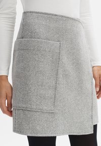 Graue gestreifte Schürze mit großer Fronttasche, gefertigt aus einem strukturierten Stoff. Der Saum ist gerade, und das Design ist minimalistisch.