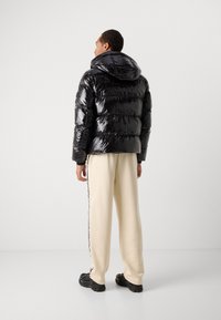Giacca a vento nera con texture lucida e trapuntata, dotata di cappuccio, abbinata a pantaloni sportivi di un crema chiaro con strisce laterali e sneakers nere.