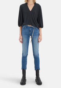 Haut en velours noir avec un col en V et des manches bouffantes. Porté avec un jean skinny bleu clair et des bottines noires. Ceinture chaîne dorée en accent.