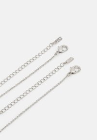 Silberne Metallkette mit drei Längen, jeweils mit runden Gliedern. Enthält Karabinerhaken und kleine Anhänger zur Befestigung.