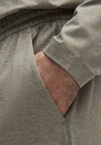 Lys grøn, tekstureret stof med et subtilt mønster; har en lomme og elastiske manchetter; close-up af en hånd, der er stukket ind i beklædningen.