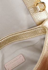 Złota, teksturowana skórzana torebka w środku z jasnym materiałem wyłożenia. Posiada złote okucie w postaci pętli i różową metkę z napisem "Coccinelle Authentic Italian Leather."
