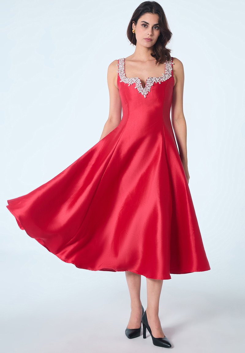 Robe en satin rouge avec une encolure en V et des bretelles ornées. Silhouette A-ligne avec un ourlet fluide, complétée par des talons hauts noirs.