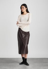 Light beige long-sleeve top with a soft texture, paired with a brown leather midi skirt. Model wears dark socks and black shoes.