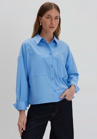Camicia azzurra chiaro con colletto, maniche lunghe e due tasche frontali. Texture liscia, vestibilità comoda e polsini delle maniche arrotolati.