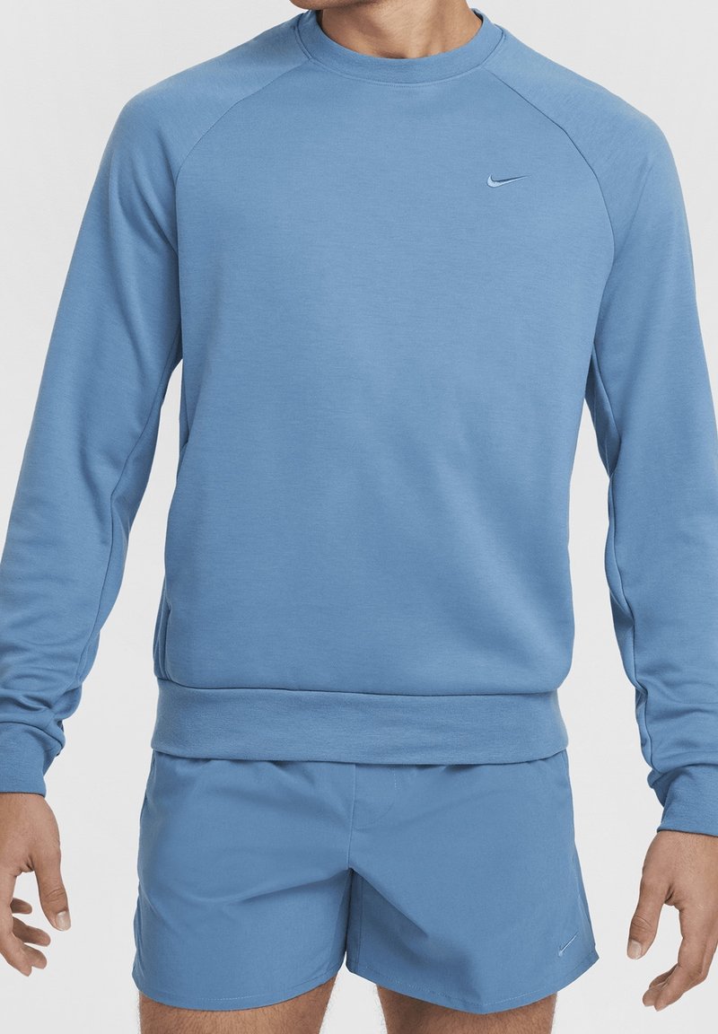 Homme portant un sweat-shirt bleu clair à manches longues et un short de sport bleu assorti avec de petits logos Nike sur la poitrine et la jambe gauche.