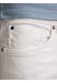 Jeans de mezclilla blanca con un bolsillo frontal que presenta un detalle de botón negro, textura suave y vista en primer plano que muestra la costura.