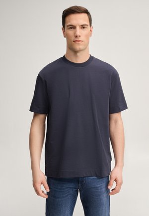 Homme debout, face vers l’avant, portant un t-shirt uni bleu foncé à manches courtes et un jean bleu, expression neutre, fond clair.