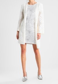 Cardigan bianco a maglia con motivo floreale sopra un vestito bianco a fantasia. Indossatrice in scarpe basse argentate, in piedi su uno sfondo neutro.