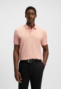 Kortärmad polo i ljusrosa bomullstyg, med klassisk krage, tre-knapps placket och subtilt broderad logotyp. Svarta byxor.