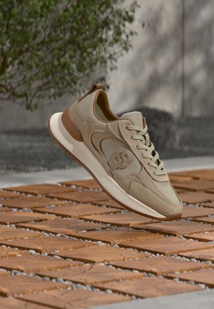 Scarpa da ginnastica beige e marrone con logo sul lato, lacci legati, che fluttua sopra un pavimento esterno in piastrelle di legno con sfondo di fogliame verde.