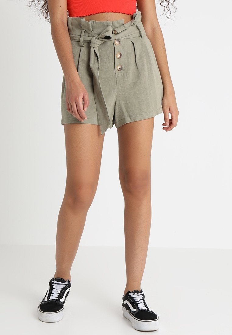 Personne portant un short taille haute vert olive avec ceinture à nouer, un crop top rouge et des baskets noires, debout devant un fond uni.