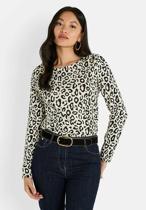 T-shirt à manches longues en crème avec un imprimé léopard noir, présentant un col rond et un détail de bouton doré à l'épaule. Porté avec un jean sombre.