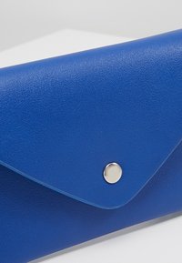 Modrá texturovaná kožená peněženka ve stylu obálky se stříbrným patentem na bílém povrchu.