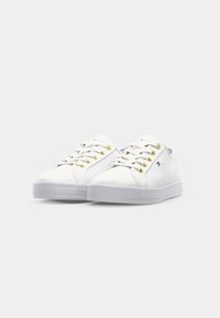 White low-top sneakers with gold eyelets and white laces, featuring a small Tommy Hilfiger flag logo on the side.