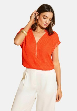 Žena v oranžovém texturovaném krátkém rukávu s předním zipem a bílých širokých kalhotách, doplněná zlatým náramkem a náušnicemi.