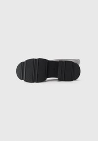 Semelle en caoutchouc noir d'une chaussure de couleur claire avec un motif texturé et une semelle striée, posée à plat sur un fond blanc.