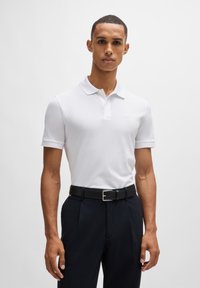 Hvit kortermet polo med klassisk krage, kombinert med mørke bukser og et svart belte med sølvspenne. Enkelt design.