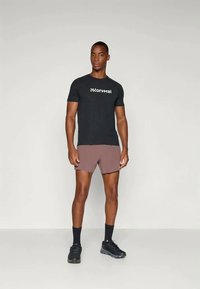 Schwarzes T-Shirt mit kurzen Ärmeln und weißem Logo, kombiniert mit braunen Sportshorts. Das Model trägt schwarze Socken und Sportschuhe.