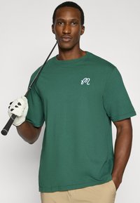 Πράσινο βαμβακερό T-shirt με κοντά μανίκια και στρογγυλή λαιμόκοψη, με μικρό λευκό κεντητό λογότυπο στο στήθος. Μπεζ παντελόνια και ένα γκολφ κλαμπ.