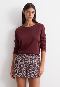 Bordeaux langemouwenshirt met logo aan de voorkant, gecombineerd met zwarte shorts met bloemmotief met roze en rode bloemen, voorzien van een elastische tailleband.