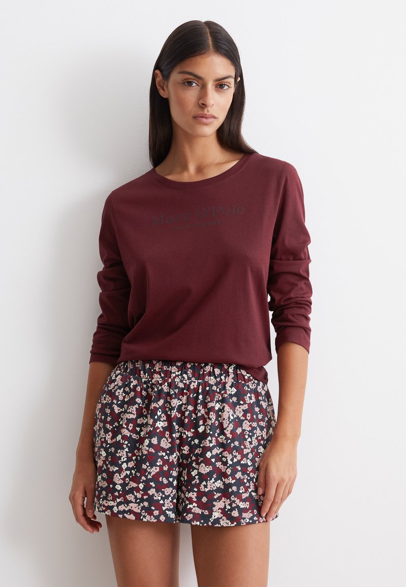 Bordeaux langemouwenshirt met logo aan de voorkant, gecombineerd met zwarte shorts met bloemmotief met roze en rode bloemen, voorzien van een elastische tailleband.