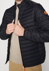 Schwarze Steppjacke mit horizontalen Abnähern, Reißverschluss vorne und hochgeschlossenem Kragen. Mit orangefarbenem Logo-Patch am linken Ärmel und strukturiertem Stoff.