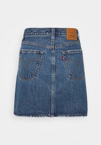 Mini-jupe en denim bleu avec deux poches arrière, surpiqûres jaunes, un patch en cuir de la marque et une petite étiquette en tissu rouge sur la poche droite.