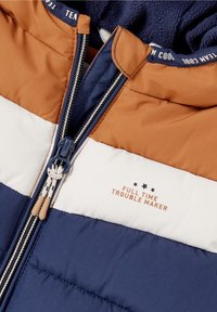 Brązowa, biała i granatowa kurtka ocieplana z zamkiem i napisem "PRAWIDŁOWY KŁOPOTLIWY" pod trzema czarnymi gwiazdami na klatce piersiowej.