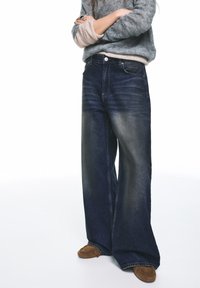 Weite Jeans aus dunklem Denim mit verwaschenem Look, hoher Taille und klassischem Fünf-Taschen-Design. Kombiniert mit braunen Mokassins.