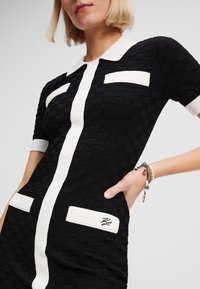 Black textured dress with short sleeves, white collar, vertical white stripe, and two white patch pockets. Annotation on the right pocket.