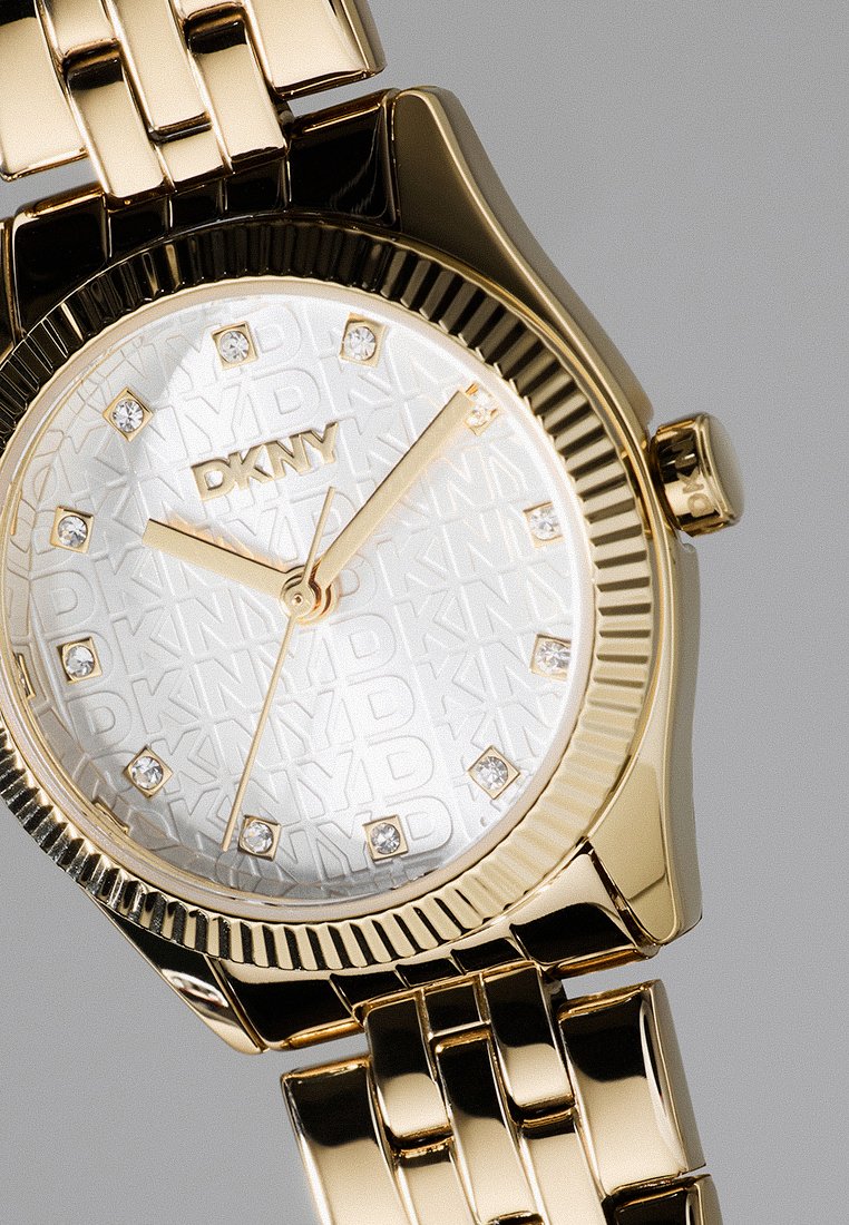 Reloj de pulsera DKNY en dorado con marcadores de hora de cristal, fondo texturizado con el logo y una correa de metal con eslabones, sobre un fondo gris claro.