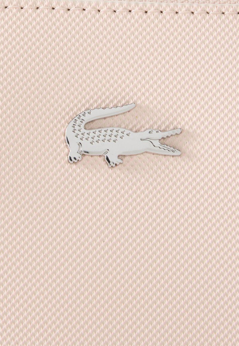 Silberne Krokodil-Logo-Nadel auf blassrosa strukturiertem Stoff mit sichtbaren Nähten am oberen Rand.