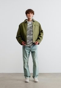 Νεαρό άτομο που φοράει πράσινο μπουφάν πάνω από γκρι παραλλαγής πουκάμισο, ανοιχτόχρωμο τζιν και λευκά αθλητικά παπούτσια, στέκεται μπροστά σε απλό λευκό φόντο.