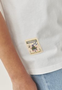 Hvid kortærmet skjorte med et dekorativt broderet patch med blomsterdesign i blå, pink og grøn, placeret på venstre side.