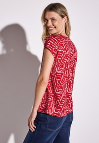 Top rosso a maniche corte con un motivo astratto bianco. Tessuto liscio, vestibilità comoda, scollatura tonda e spacchi laterali all'orlo.