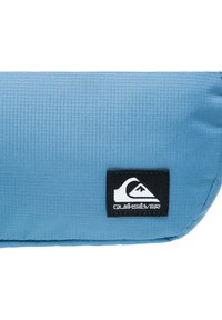 Blauer Stoffbeutel mit einer strukturierten Oberfläche und einem schwarzen Quiksilver-Logo-Patch. Kompakte rechteckige Form, strapazierfähiges Material und dezente Nähte.