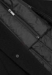 Wnętrze czarnego płaszcza z pikowanym podszyciem i dwoma kieszeniami na zamek. Posiada matowe wykończenie oraz pojedyncze zapięcie na guzik na zewnętrznym materiale.