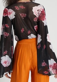 Blusa nera trasparente con stampa floreale in tonalità di rosa e rosso, caratterizzata da ampie maniche a campana, abbinata a pantaloni a vita alta a pieghe arancioni.