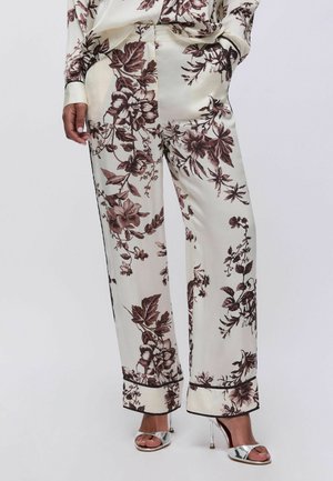 Pantaloni ampi in seta color crema con stampa floreale marrone, dettagli in piping nero, abbinati a sandali argento con tacco alto e punta aperta.