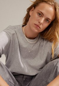 Grå bomulds t-shirt med afslappet pasform, kombineret med lagdelte sølv halskæder og grå jeans. Blød tekstur og minimalistisk design.