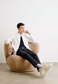 Una poltrona rotonda, beige e texturizzata; una persona vestita con una camicia bianca e pantaloni neri siede comodamente, indossando delle sneakers, su uno sfondo minimalista bianco.