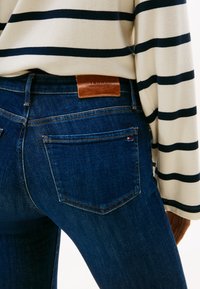 Rückenansicht einer Person, die dunkelblaue Jeans mit Tommy Hilfiger Lederlabel und einen cremefarbenen Pullover mit schwarzen horizontalen Streifen trägt.