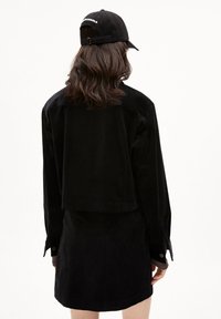 Czarny strój z korduroyu, składający się z kurtki o skróconym kroju i pasującej spódnicy. Kurtka ma długie rękawy, mankiety na guziki oraz gładką teksturę.