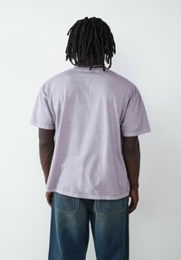 T-shirt z krótkim rękawem w kolorze lawendowym, wykonany z bawełny, o luźnym kroju, z widocznym szwem i okrągłym dekoltem, noszony z niebieskimi dżinsami.