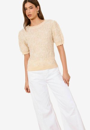 Femeie purtând un top crem din dantelă cu mâneci scurte și modele florale, precum și pantaloni albi cu picioare largi, stând cu o mână relaxată pe lângă corp.