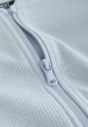 Világoskék cipzáras dzseki texturált anyagból. Simított cipzárral és bordázott részletekkel rendelkezik, kiemelve a modern, funkcionális dizájnt.